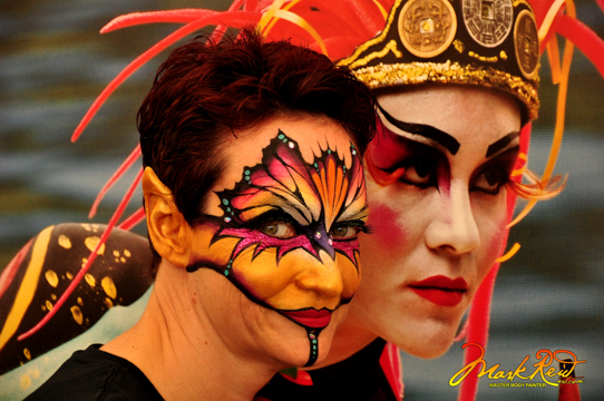 woman in a painting on the upper part of her face with gold highlights and elf ears in front of an asian themed poster